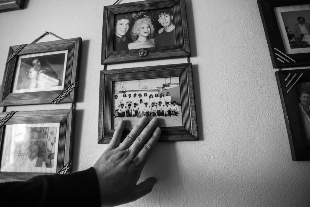 A hand touches a framed black-and-white photo of a group on a wall, surrounded by other framed family and group photos. | En-dash.co.uk